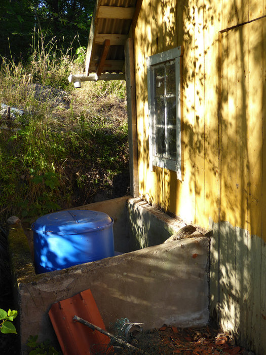 Hörn av gul vedbod med synlig skadad grundmur och en blå tunna vid husväggen.