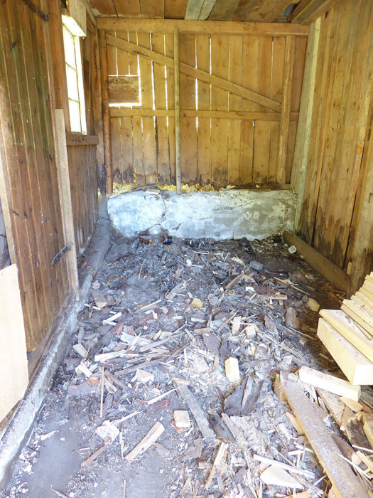 Vedbod interiör efter rensning med skadad grundmur och spån på golvet.