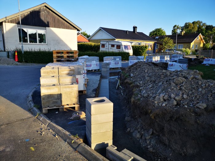 Pågående arbete med stenmur, betongblock staplade på pallar och en grushög nära en vägkant.