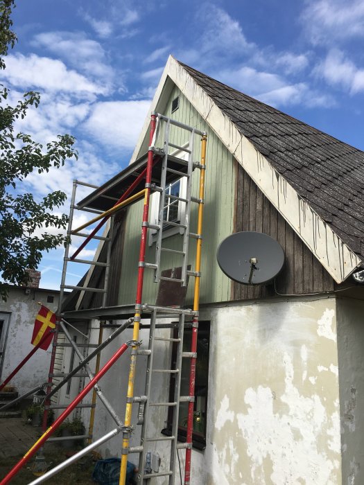 Byggställning framför gaveln på ett hus under renovering med nymålad panel och gammal satellitdisk.