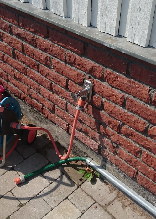 Ny Gustavsberg utekran monterad på sprucken tegelvägg vid träplank, med anslutna vattenslangar.