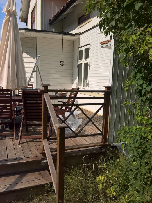 Trädäck med trädgårdsmöbler och en stängd parasoll, stegar lutade mot ett hus, omringat av grönska. Målning pågår.