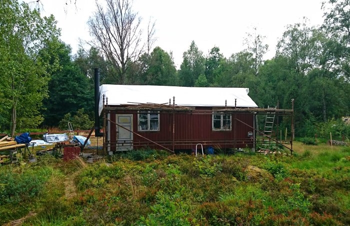 Renovering av hus med ny råspont och papp på tak, uppsatt byggställning och omgivande natur.