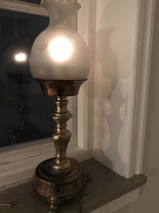 Gammaldags bordslampa med tänd glödlampa på ett fönsterbräde som ger ett varmt ljus och skapar skuggspel på väggen.