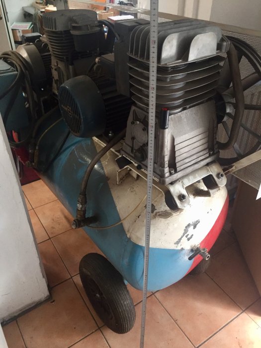 En kompressor med blå och röd tank och flera motorer i en verkstadsmiljö med tillhörande slangar och mätskala.