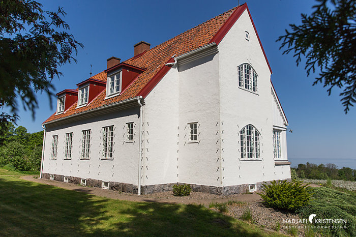 Hus från 1912 med utstickande armering på vit fasad under blå himmel, möjlig spaljé för växter.