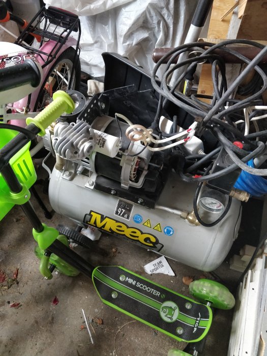 Rörigt garage med en gammal luftkompressor märkt 'Meec' och en barnsparkcykel 'MINI SCOOTER' på golvet.