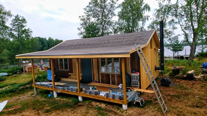 Nybyggd stuga med råspont och papp på taket, veranda väntar på ytpapp, omgiven av natur.