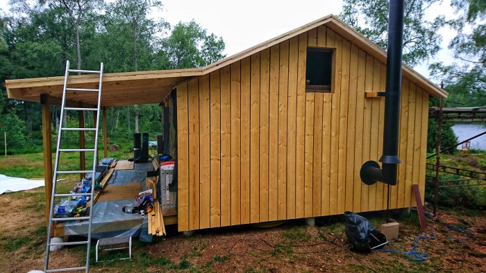 Halvfärdigt trähus med råspont och papp på norrsidan, verandan under tak och verktyg framför.