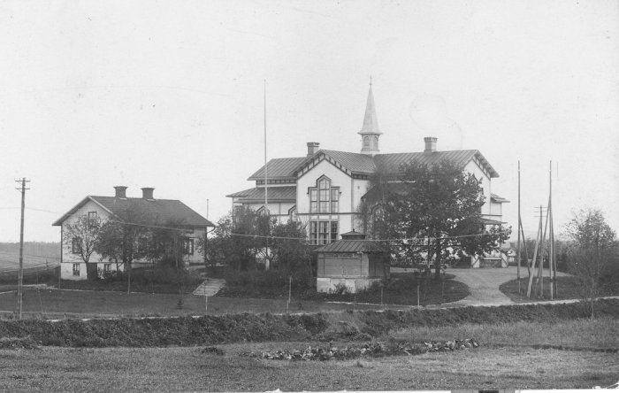 Svartvit bild av ett gammalt herrgårdsliknande hus med torn och flera skorstenar, i en lummig trädgård.