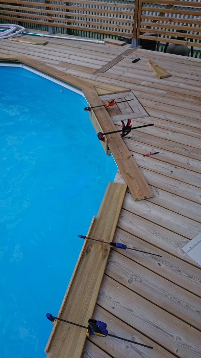 Trädäck under konstruktion runt en pool med träbitar, tvingar och verktyg.