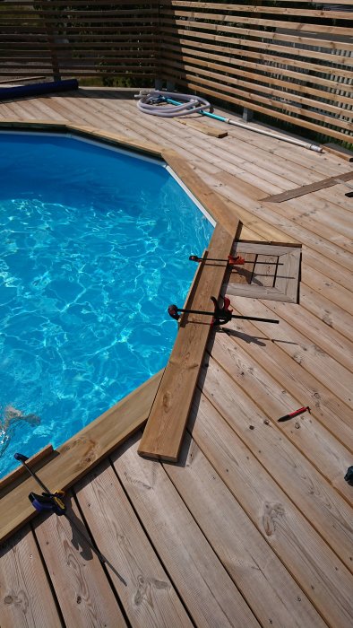 Trädäck runt pool med en jigg av trä och masonit fastklämd med tvingar, virke och verktyg synliga.