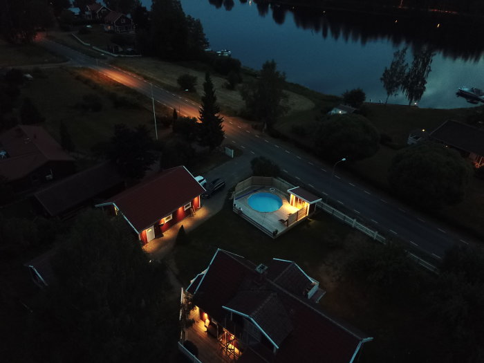 Kvällsvy över en trädgård med upplyst pool omgiven av däck och hus, intill en väg och vatten.