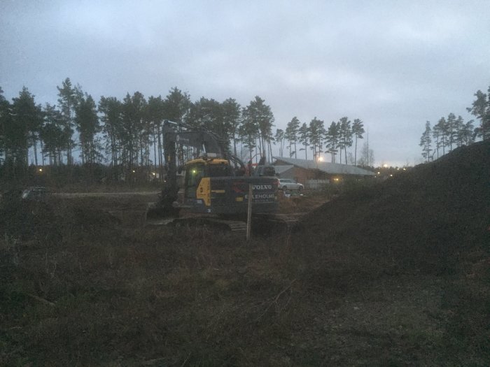 Grävmaskin som schaktar en tomt i skymningen med sandhögar och träd i bakgrunden.