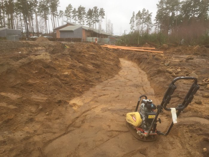 Uppschaktad tomt med sandfyllda rännor och grävmaskin, i bakgrunden syns hus och träd.