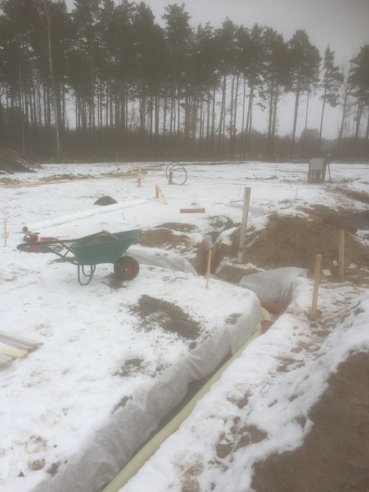 Byggplats med dränering och dagvattenledningar under arbete, snötäckt mark med träd i bakgrunden.