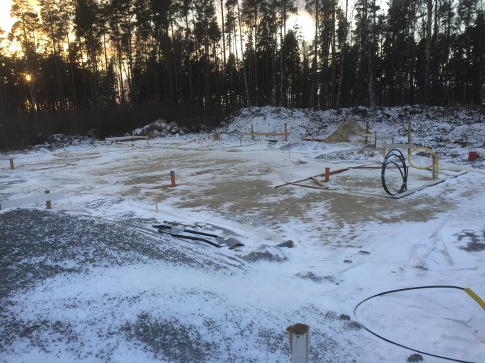 Byggplats med markerade ramar för avlopp i sand och stuprör, delvis täckt av snö.