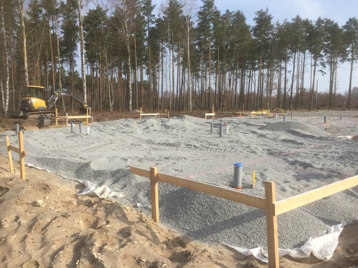 Grundarbete med marduk och makadam på byggplats, grävmaskin i bakgrunden och skog runtomkring.