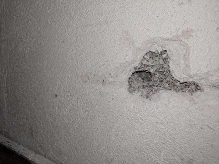 Hål i putsad vägg som avslöjar mörka träflisor och stålnät, misstänkt betonghålsten bakom.