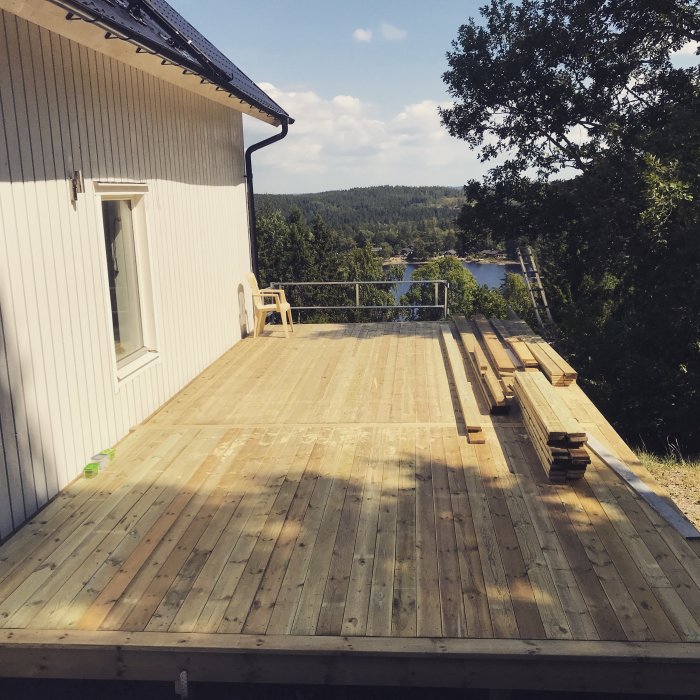 Nyinstallerad trätrall på altan vid vit husvägg, med staplade trallbrädor på sidan och sjöutsikt.