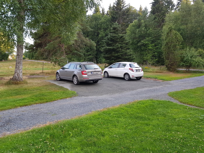 Två bilar parkerade på nygjord grusparkering bredvid en gräsmatta och träd.