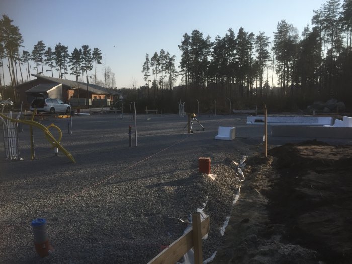 Byggplats med makadamgrund och synliga rörledningar, i bakgrunden skog och byggnader.