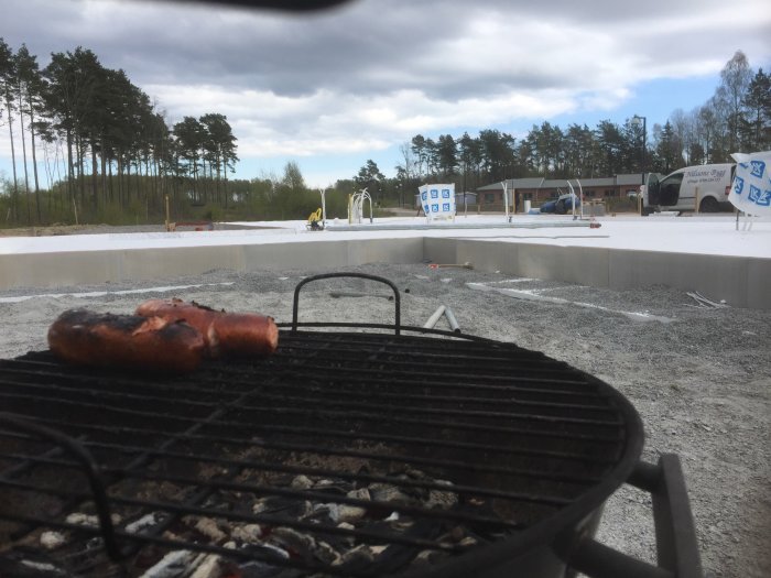 Grundarbete syns i bakgrunden med S100 cellplast, och en korv grillas i förgrunden på ett bygge.