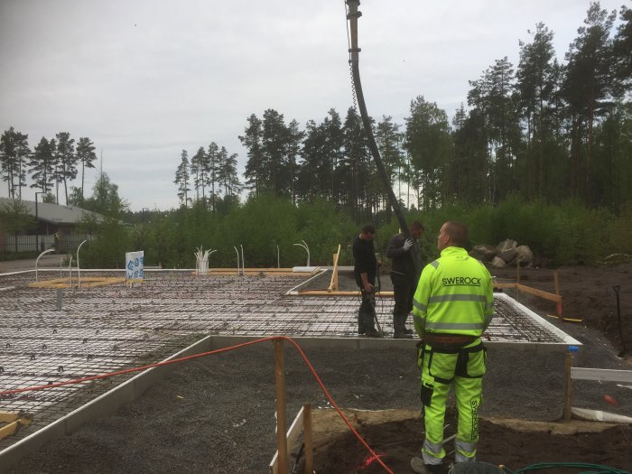 Arbetare i säkerhetskläder gjuter en betongplatta, med byggmaterial och skog i bakgrunden.