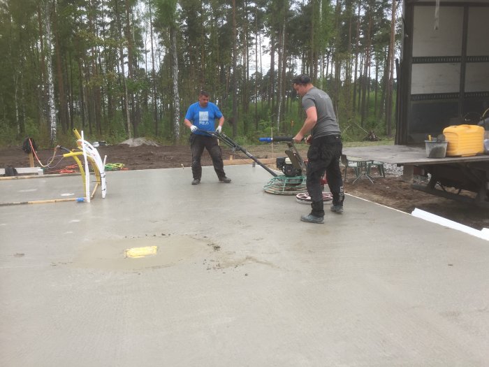 Två personer glättar nyligen gjuten betongplatta utomhus, med skog och byggmaterial i bakgrunden.