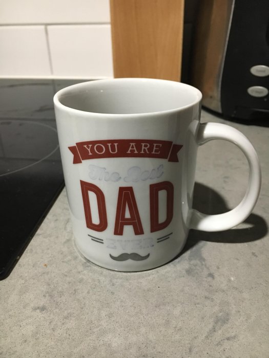 Vit mugg med text "YOU ARE THE BEST DAD EVER" på ett köksbord.