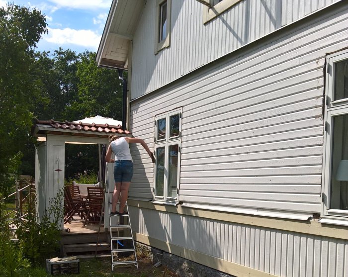 Person på stege förbereder husvägg för målning under solig dag.