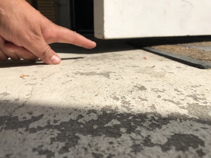 Någons hand pekar på en sliten och fläckig betongförstubro med skrapmärken och sprickor vid en dörrtröskel.