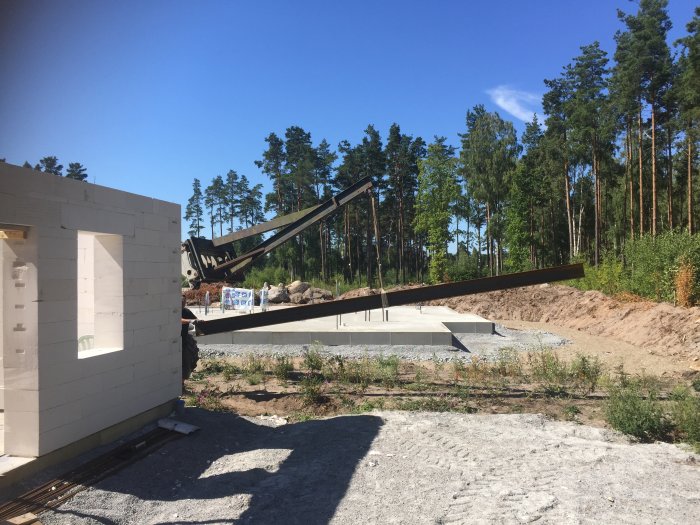 Järnbalk lyfts av kran över byggarbetsplats med stödpelare och delvis byggt hus.