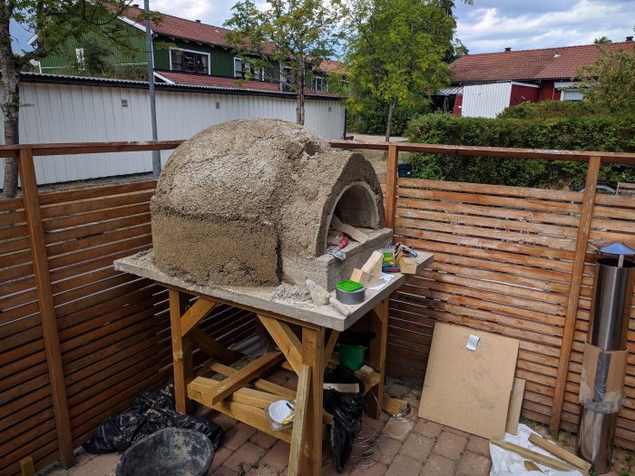 Ofärdig hemmabyggd pizzaugn i vermicrete och isolering på en träbänk utomhus med byggmaterial runtomkring.