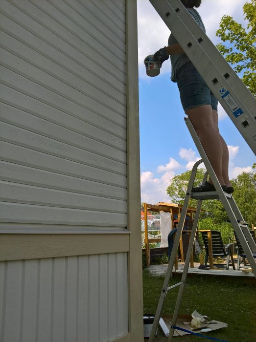 Person på stege som målar en vägg på hus under en klar himmel, byggmaterial synligt på marken