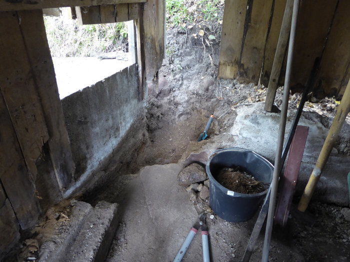 Urgrävd del av en vedbod med verktyg, en svart hink med jord och synlig gammal grund.