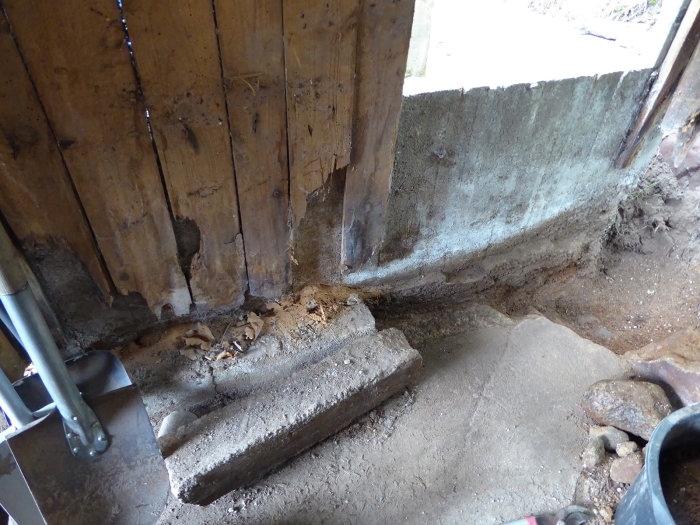 Rensad hörna i vedbod med betongklack och urgrävt golv inför gjutning, bredvid trävägg och skottkärra.