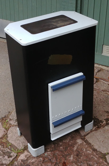 Porin Matti braskamin i svart med lite stenskador på fronten, vit med blå detaljer, placerad utomhus.