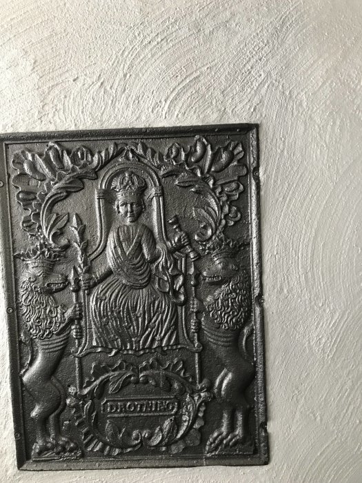 Dekorativ svart metallplakett med relief av en figur och lejon på en putsad vägg i betonggrå nyans.