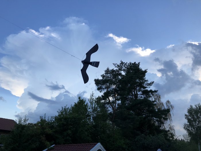 Svart fågelskrämma liknande en fågel flyger mot molnig himmel omgiven av träd.