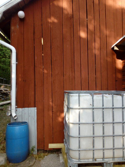 IBC-tank bredvid röd trävägg med stuprör och blå regntunna, grovkapat virke synligt under tanken.