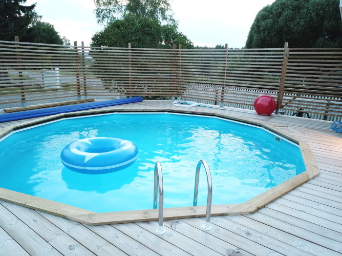 Rund ovanmarkspool med blått vatten omgiven av träterrass, fris mot poolen synlig, uppblåsbar ring och röd boll i poolen.