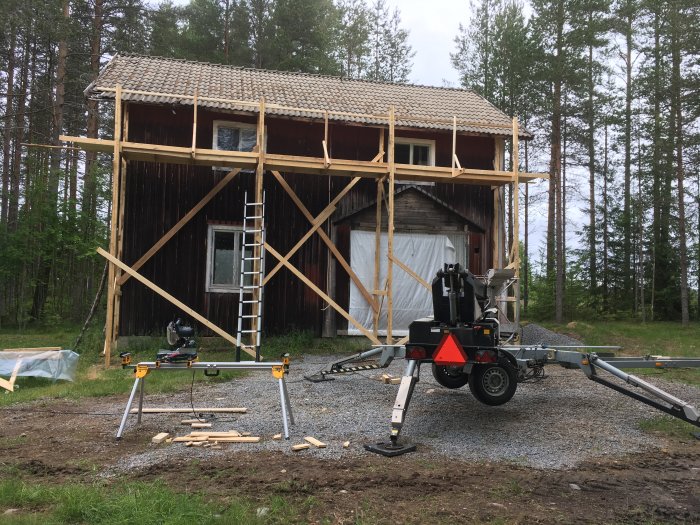 Träställning byggd runt rödfärgad ladugård med betongpannetak, byggarbetsplats med verktyg i förgrunden.