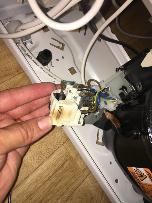 Hand håller startrelä till Elektro Helios frys på vit bakgrund med verktyg och kablar.