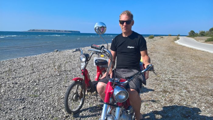 Person i Bygga Hus-tröja på röd moped vid strand med hav och klippa i bakgrunden.