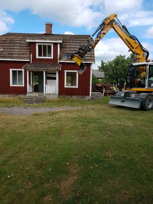 En grävmaskin framför ett rött timmerhus under rivning, med rivningsavfall på marken.