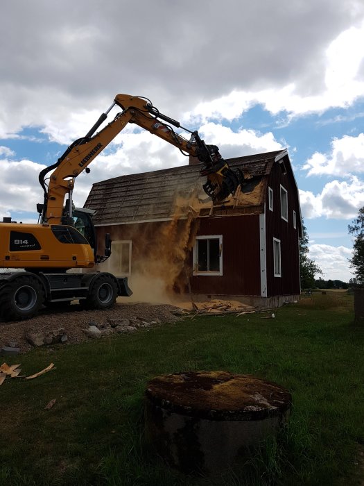 Gul grävskopa river taket på ett rött trähus, med en stofthög och dammoln runtomkring.