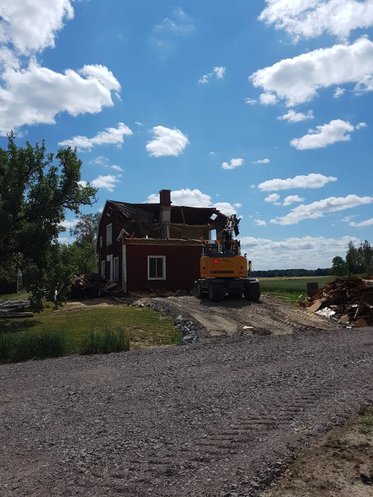 Grävmaskin vid delvis rivet rött hus med spillror runtomkring under klarblå himmel.