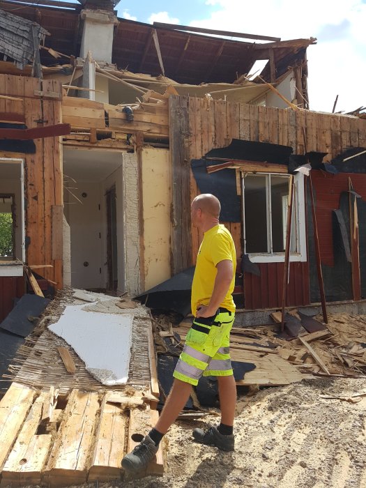 Person i arbetskläder framför halvt nedrivet hus med bråte runtomkring.