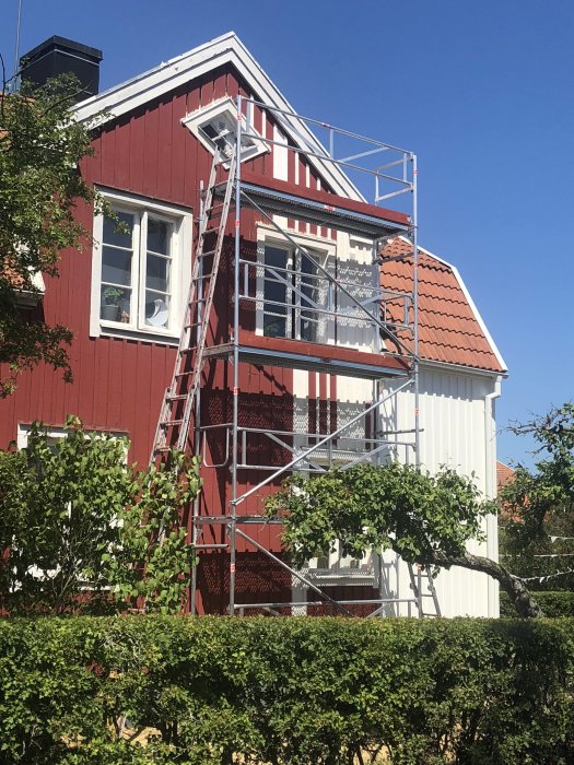 Rött trähus med uppsatt byggställning längs ena väggen och grönskande häck i förgrunden.